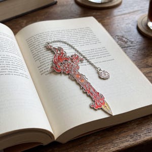 Może przedstawiać: Metalowa zakładka do książki w kształcie miecza z motywem płomieni w odcieniach czerwieni, pomarańczu i żółci. Miecz posiada misternie zdobione srebrne detale i jest przymocowany do srebrnego łańcuszka z małą, okrągłą zawieszką w kształcie spirali.