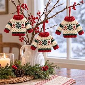 May include: Three miniature crocheted sweater ornaments featuring a cream-colored base with red and green snowflake patterns, red ribbed collars, cuffs, and hems. Each sweater has a red yarn loop at the top for hanging on branches with red berries.