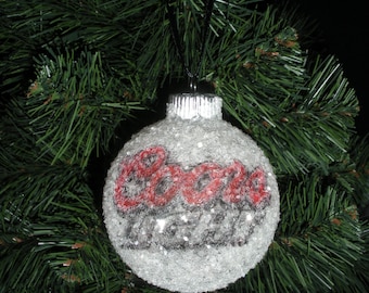 May include: A round, silver Christmas ornament with the word "Coors" in red script and black accents. The ornament is covered in glitter and hangs from a black ribbon against a backdrop of green Christmas tree branches.