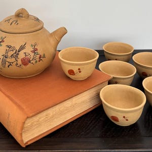 May include: A tan ceramic teapot with a bamboo-shaped handle and a lid, decorated with hand-painted birds and floral motifs in red, green, and black. Five matching small tan ceramic tea cups are arranged nearby, each featuring a single painted red flower.
