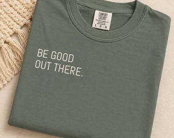 May include: A sage green crew neck t-shirt featuring the white text BE GOOD OUT THERE printed on the front. The shirt has a Comfort Colors brand label on the inside collar indicating a size large, made of ring-spun cotton in Honduras.