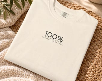May include: A cream-coloured short-sleeve t-shirt featuring the black text 100% SUBJECT TO CHANGE. printed on the chest. The shirt has a crew neckline and a Comfort Colors brand label visible on the inside of the collar area near the neck.