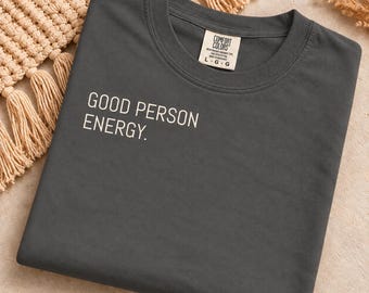 May include: A charcoal grey crew neck t-shirt featuring the white text GOOD PERSON ENERGY. printed on the left chest area. The shirt has a Comfort Colors brand label inside the collar indicating a size large, made in Honduras of 100% cotton.