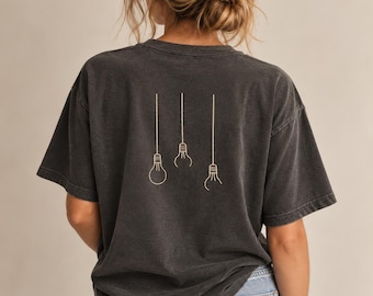 May include: A charcoal grey short-sleeve t-shirt featuring a minimalist line art graphic on the back. The design consists of three light bulbs hanging from thin vertical lines, rendered in a light beige colour against the dark fabric background.