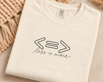 May include: A cream-coloured short-sleeve t-shirt featuring a black graphic design on the chest. The design includes mathematical symbols for less than, equal to, and greater than, with the cursive text "less is more" printed directly underneath.