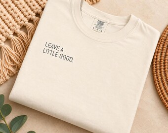 May include: A cream-coloured short-sleeve t-shirt with a crew neckline. The shirt features the black text "LEAVE A LITTLE GOOD." printed on the upper left chest area. The interior neck label displays the brand name Comfort Colors and size L.
