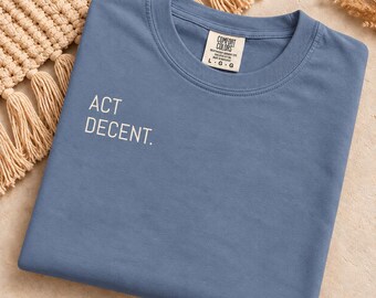 May include: A blue crew neck t-shirt featuring the white text ACT DECENT. printed on the left chest area. The shirt includes a Comfort Colors brand label at the inner neckline indicating a size large, made in Honduras, and 100% ring spun cotton.