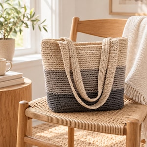 Puede incluir: Un bolso tote rectangular de ganchillo con un diseño de bloques de color con tres franjas horizontales en crema, gris claro y gris carbón oscuro. El bolso tiene dos asas de hombro de ganchillo a juego en color crema.
