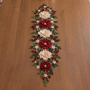 May include: A long, narrow crochet table runner featuring a repeating pattern of large, layered cream and deep red flowers. The floral design is accented with small white blossoms, dark green leaves, and clusters of tiny red berries throughout.