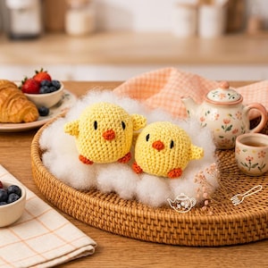 May include: Two round, yellow crocheted chicks with orange beaks and feet, black eyes, and small wings, resting on a bed of white polyester fiberfill inside a round, woven rattan tray, accompanied by a floral teapot and a small matching teacup.