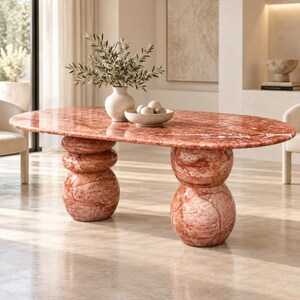 Peut inclure: Une table à manger ovale en marbre rouge poli avec des veines blanches marquées. La table repose sur deux bases imposantes, chacune composée de deux formes sphériques arrondies empilées. Un vase et un bol en céramique sont posés dessus.