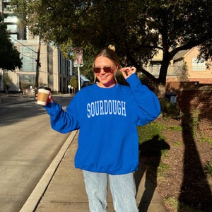 May include: A bright blue crewneck sweatshirt featuring the word SOURDOUGH printed in white capital letters across the chest. The garment has long sleeves and a ribbed collar, cuffs, and hem, paired with light blue denim jeans.