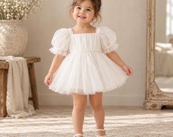 Puede incluir: Un vestido blanco con mangas cortas abullonadas y una falda de tul en capas. El corpiño está fruncido con un escote cuadrado, y el vestido se combina con zapatos planos blancos a juego con un pequeño lazo en la parte delantera.