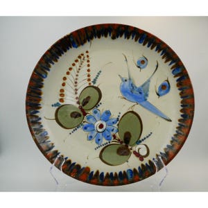 May include: A round ceramic plate with an off-white base featuring a hand-painted blue bird, a central blue flower, and green leaf motifs. The rim is decorated with a repeating pattern of dark brown and blue brushstrokes.