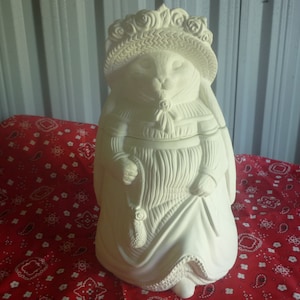 Puede incluir: Figura de cerámica de un conejo vestido con un gorro y un vestido, sosteniendo un bolso. La escultura blanquecina tiene texturas detalladas y está sobre un fondo rojo estampado.