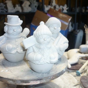 May include: Three unpainted ceramic snowman figurines. One wears a top hat and holds a broom. Another wears a knit cap and holds a sign. The third snowman wears a knit cap and scarf.