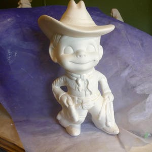 May include: A ceramic figurine of a smiling cowboy boy, wearing a cowboy hat, fringed shirt, and pants. The figurine is painted in a solid white color and is posed with one hand on his hip and the other holding a sack.