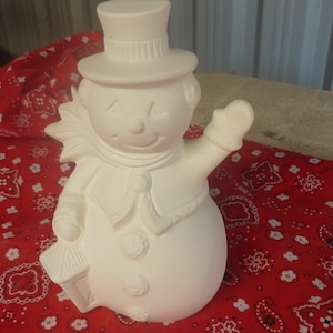 Może przedstawiać: Niemalowana ceramiczna figurka bałwana z cylindrem, szalikiem i rękawicami. Bałwan trzyma latarnię i macha. Figurka stoi na czerwonej tkaninie we wzory.