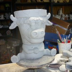 May include: A light gray ceramic planter shaped like a cartoon cow. The planter has a wide opening, a detailed cow face, and sits on a round base. The cow is in a seated position with its legs extended.