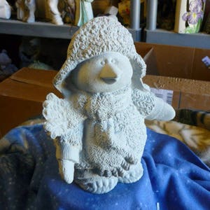 May include: A white ceramic penguin figurine with a fluffy hat and scarf. The penguin is standing on a blue blanket.