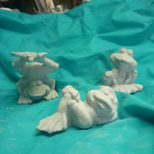 Könnte beinhalten: Drei weiße Keramik-Froschfiguren auf einem türkisfarbenen Hintergrund. Ein Frosch hält ein Blatt, ein anderer sitzt mit gekreuzten Beinen und der dritte liegt auf dem Rücken mit den Beinen in der Luft.