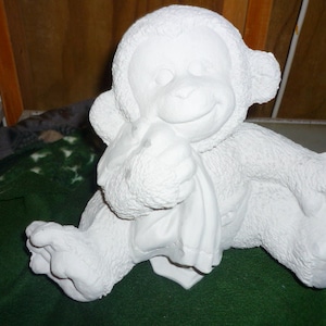 Pode incluir: Figura de macaco de cerâmica branca com uma expressão brincalhona, segurando um cobertor em suas mãos. O macaco está sentado em uma superfície verde.