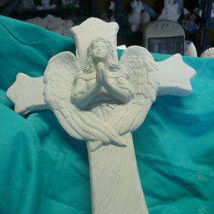 May include: A white, cross-shaped sculpture featuring an angel with outstretched wings and clasped hands. The angel is centered on the cross, which has star-shaped ends. The sculpture is set against a teal fabric background.