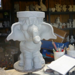 Puede incluir: Maceta de cerámica blanca en forma de elefante con una trompa grande y orejas anchas. La maceta está sentada en una plataforma giratoria y está lista para ser pintada.