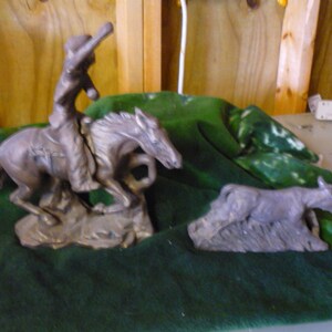 Op de afbeelding: Twee bronzen sculpturen van paarden. Een sculptuur toont een cowboy die een paard berijdt, terwijl de andere sculptuur een paard toont dat alleen staat. De sculpturen worden tentoongesteld op een groene stoffen ondergrond.