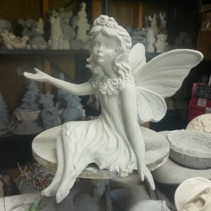 May include: A white ceramic fairy figurine with large wings, seated on a round base. The fairy is wearing a dress and a floral crown, with one arm extended. Other ceramic figurines are visible in the background.