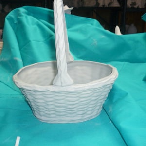 Puede incluir: Una cesta de cerámica blanca con un diseño tejido y un asa. La cesta está sobre una superficie de tela turquesa.