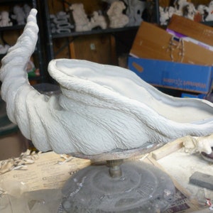 May include: A white ceramic planter shaped like a curled horn. The planter is on a rotating stand and has a textured surface.