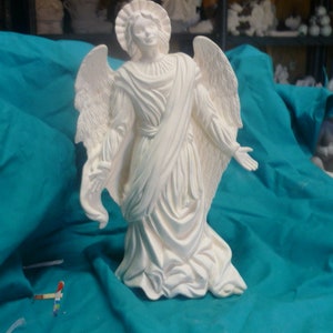 May include: White ceramic angel figurine with wings outstretched and a flowing robe. The angel is standing on a teal blue surface.