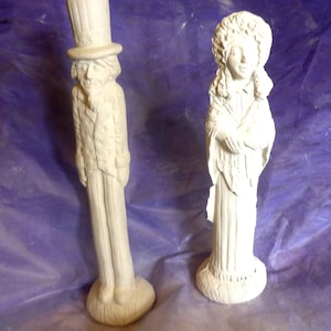 Könnte beinhalten: Zwei weiße Keramikfiguren. Eine ist eine hohe, schlanke Figur eines Mannes mit Zylinder und Anzug. Die andere ist eine Frau in einem langen Kleid und Schal. Beide Figuren haben detaillierte Gesichtszüge und stehen auf runden Sockeln.