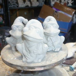 Könnte beinhalten: Drei weiße Keramik-Froschfiguren mit glasierter Oberfläche. Die Frösche sitzen auf einem runden Holzsockel.