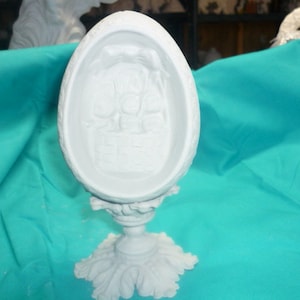 May include: White ceramic egg-shaped sculpture with a detailed design on the front. The sculpture is on a pedestal with a decorative base.