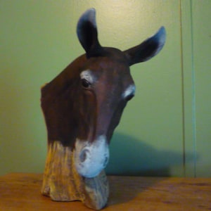 May include: A brown wooden sculpture of a mule's head with a white nose and black eyes. The sculpture is mounted on a wooden base.