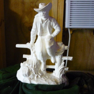Pode incluir: Figurine de cerâmica branca de um cowboy sentado em uma cerca. O cowboy está usando um chapéu e tem uma pose relaxada.