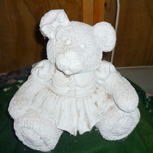 Puede incluir: Figurilla de cerámica blanca de un oso de peluche con un vestido blanco y un lazo en la cabeza. El oso está sentado sobre una superficie verde.