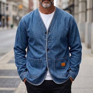 May include: A blue denim V-neck cardigan jacket featuring a button-front closure, long sleeves, and two large patch pockets on the front. A small rectangular tan leather-like patch is stitched onto the lower left pocket. The garment is worn over a plain white t-shirt.