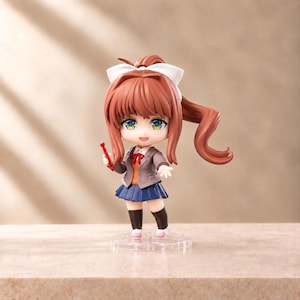 Peut inclure: Une figurine anime de style chibi représentant un personnage aux longs cheveux bruns en queue de cheval, avec un nœud blanc, des yeux verts et un uniforme scolaire composé d'un blazer gris, d'un gilet orange, d'un ruban rouge et d'une jupe bleue.