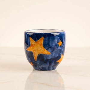 May include: A small ceramic cup featuring a deep blue background with a hand-painted, textured finish. The exterior is decorated with multiple bright orange stars of varying sizes scattered across the surface. The interior of the cup is white.