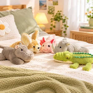 May include: A collection of plush animal toys resting on a white textured blanket, including a gray hippopotamus, a tan fennec fox, a pink axolotl, a gray rhinoceros, and a green crocodile with a serrated back ridge.