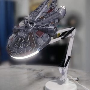 May include: A desk lamp featuring a grey and silver model of a starship as the light shade. The lamp has a white adjustable articulated arm with black springs and a round white base. A bright white light emits from the underside of the starship.