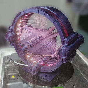 Könnte beinhalten: Eine dunkelviolette, kreisförmige, mechanische Struktur mit einem dreieckigen Raumschiffmodell in der Mitte. Der Ring wird von innen durch helles rosa Licht beleuchtet, das dünne, strahlende Linien in den Innenraum projiziert. Ein weißes Stromkabel ragt aus der Basis.