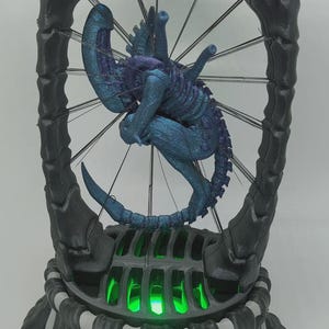 May include: A 3D printed sculpture featuring a metallic blue and purple creature suspended by thin wires within a dark, skeletal, cylindrical frame. The base of the structure is illuminated with bright green light, highlighting the intricate details.