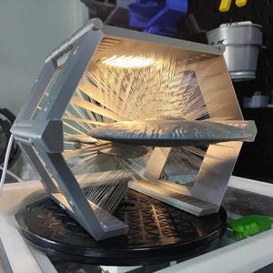 Könnte beinhalten: Ein grauer, 3D-gedruckter, sechseckiger Ausstellungsrahmen mit einer zentralen horizontalen Ablage, auf der ein untertassenförmiges Objekt ruht. Der Rahmen besteht aus dünnen, strahlenförmigen weißen Filamentlinien, die die Außenstruktur mit der Mitte verbinden.