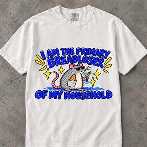 May include: A white short-sleeve t-shirt featuring a graphic of a gray rat wearing sunglasses, holding a martini glass and a loaf of bread. The text reads: I AM THE PRIMARY BREADLOSER OF MY HOUSEHOLD, surrounded by yellow star accents.