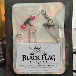 Peut inclure: Un montage de pêche dans une pochette holographique transparente avec une étiquette indiquant BLACK FLAG TACKLE HEAVY DUTY RED SNAPPER RIG 6' 130 LB MONO LEADER | 8/0 HOOK. Le montage comprend du fil monofilament, un émerillon et deux perles rouges.