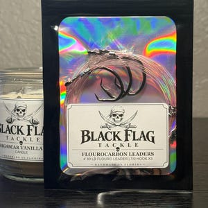 May include: A black pouch with a holographic center containing three fishing hooks attached to clear fluorocarbon leaders. The label reads BLACK FLAG TACKLE, FLOUROCARBON LEADERS, 4' 80 LB FLOURO LEADER | 7/0 HOOK X3, and HANDMADE IN FLORIDA.